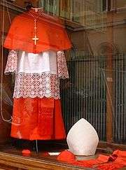 Vêtements de cardinal ; de haut en bas : mozette rouge, croix apostolique, rochet de dentelle blanche, bas de la soutane rouge; au pied du mannequin de gauche à droite: barrette en miniature, calotte, mitre (mitra simplex) et étole. 